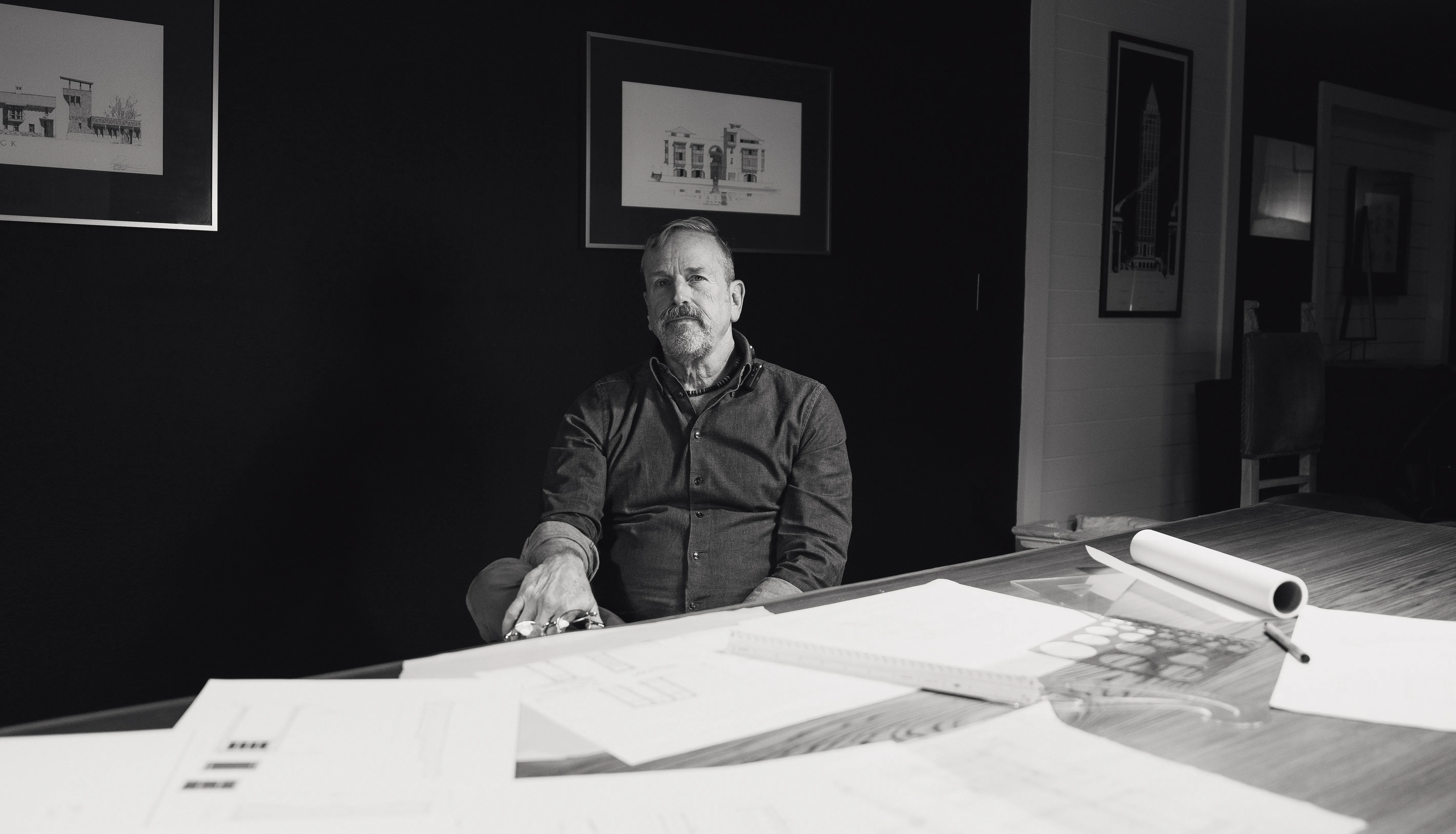 Black-and-white brand portrait of architect Lew Oliver at his desk with framed building elevations, photographed by Grit Blueprint