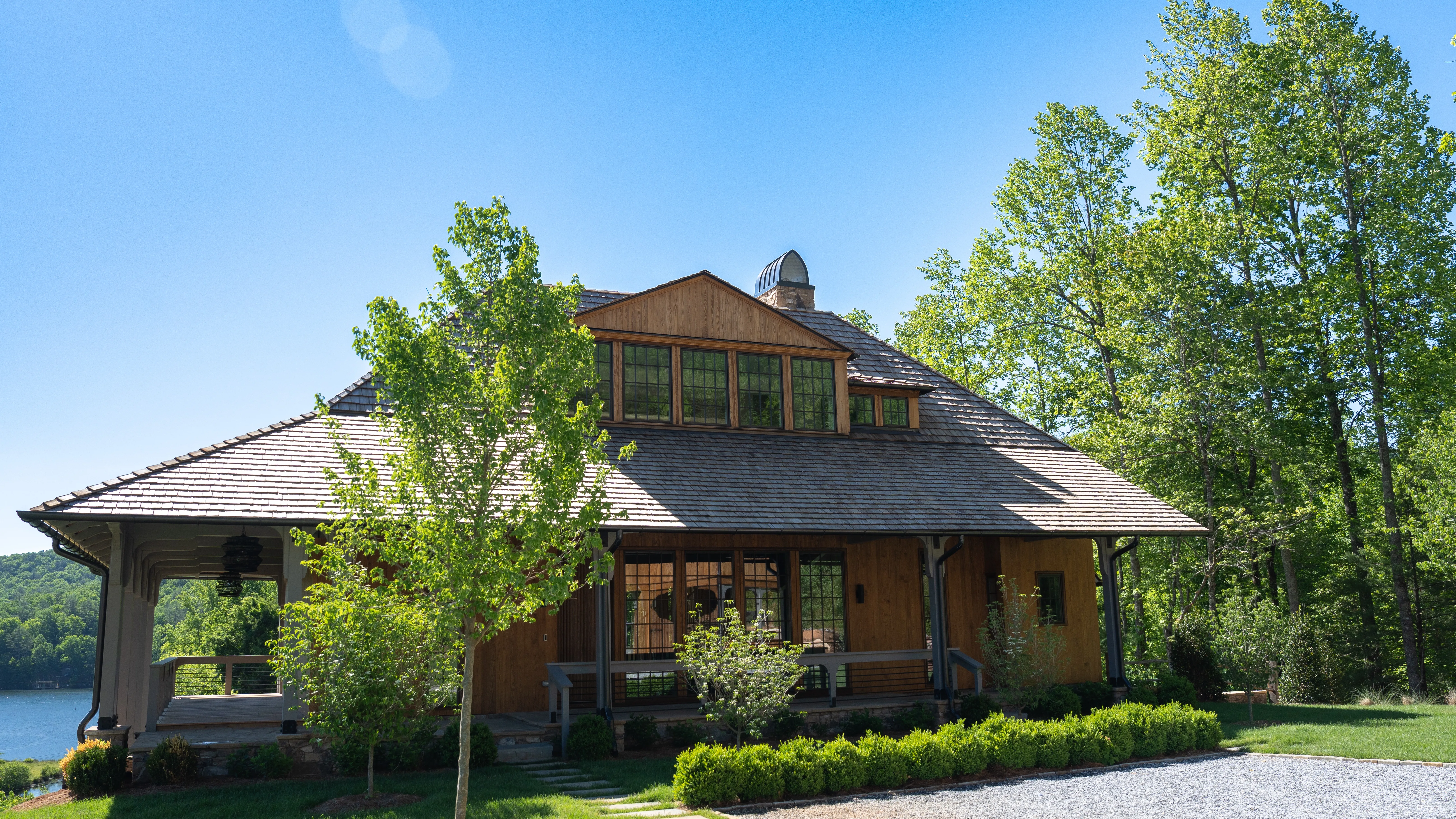 Lakeside elevation of the Burton Show Home, a cedar-shingle home with a hipped roof and wraparound porch, photographed June 2026 by Grit Blueprint