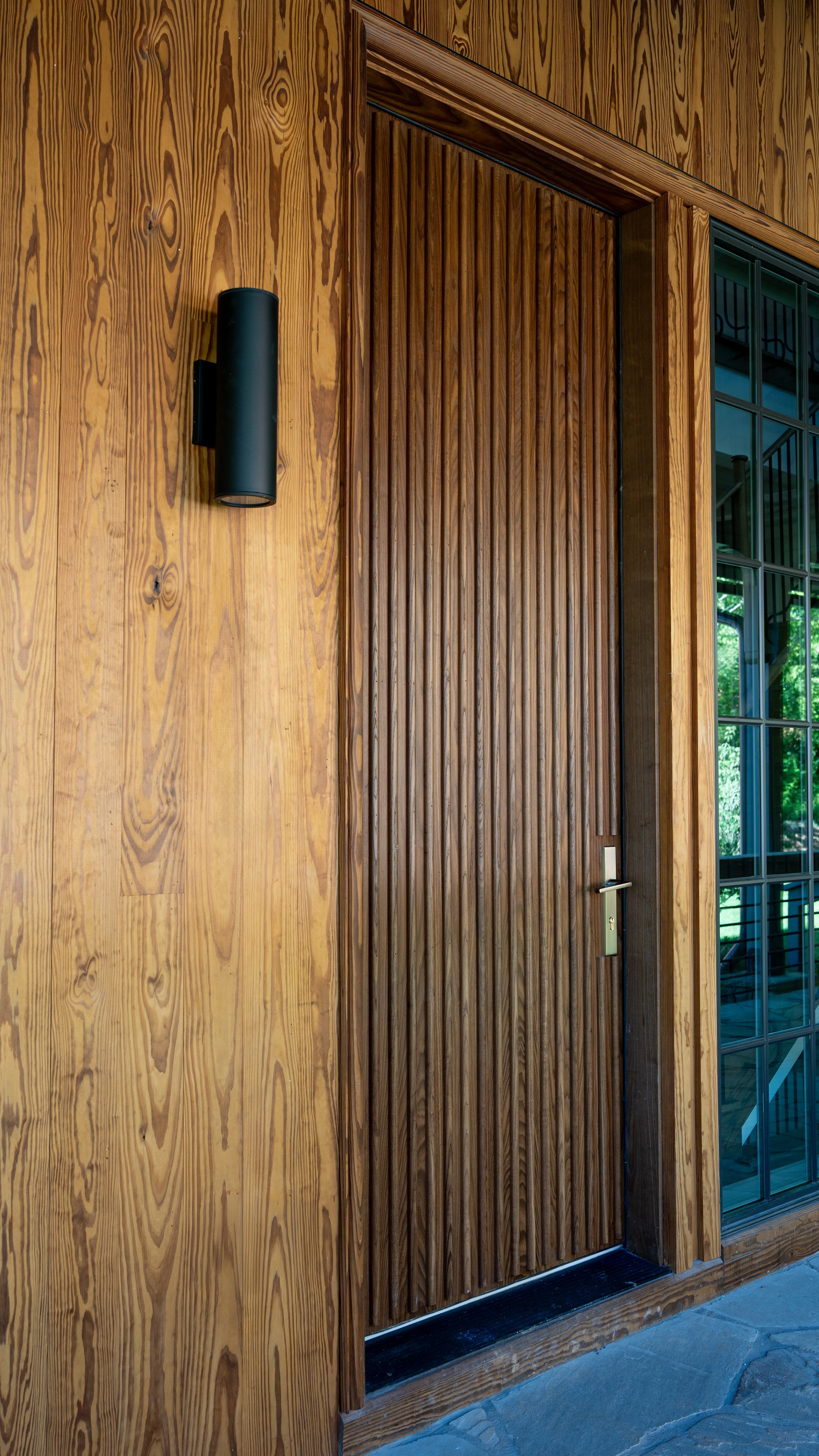 Thermally modified custom ash entry door with a reeded detail by Magbee Luxury, framed by vertical-grain pine siding and a black sconce, photographed by Grit Blueprint