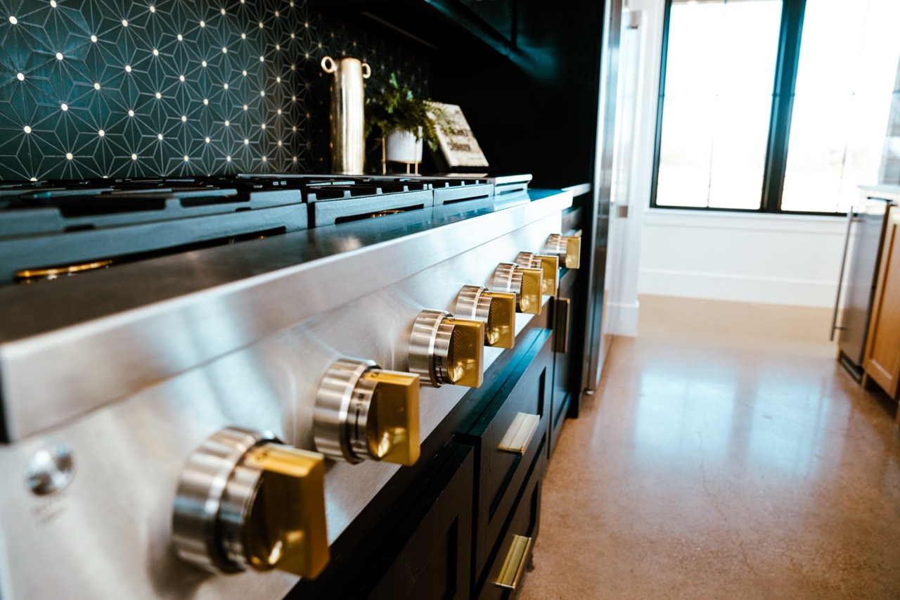 Professional range detail with brushed-steel and brass control knobs against a black geometric tile backsplash, photographed by Grit Blueprint