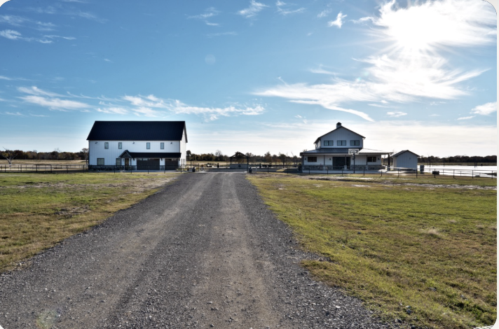 Gravel drive leading between the two white barndominium structures across the Stillwater Ranch estate under a clear Texas sky, photographed by Grit Blueprint