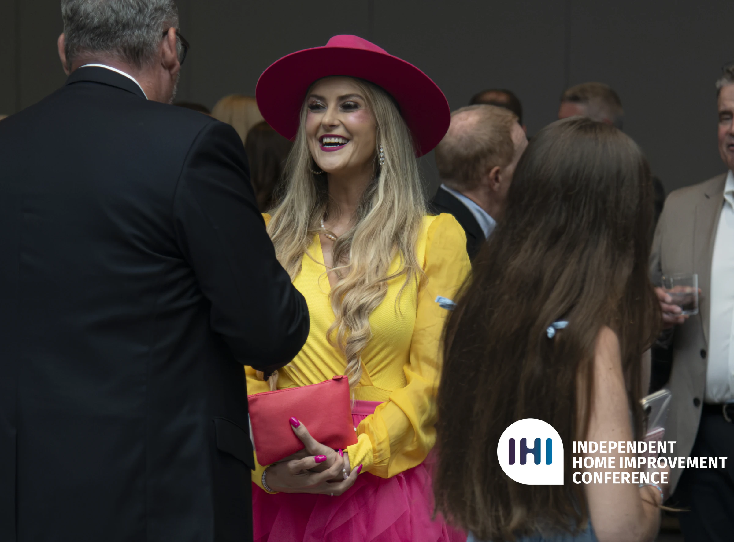 Stefanie Couch, in a pink hat and yellow top, smiles while talking with attendees at a crowded IHI conference gathering.
