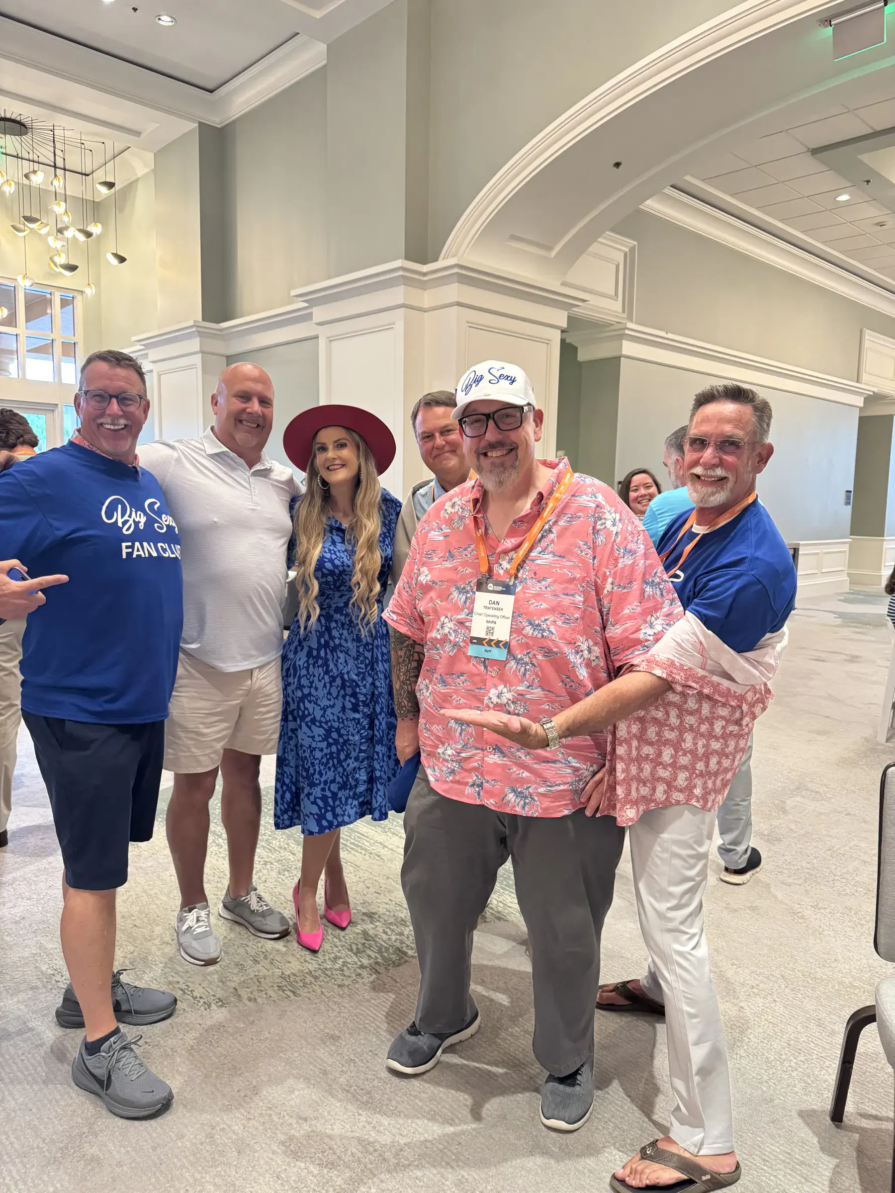 Six attendees pose together in a bright conference hall, two wearing matching Big Sexy Fan Club shirts.