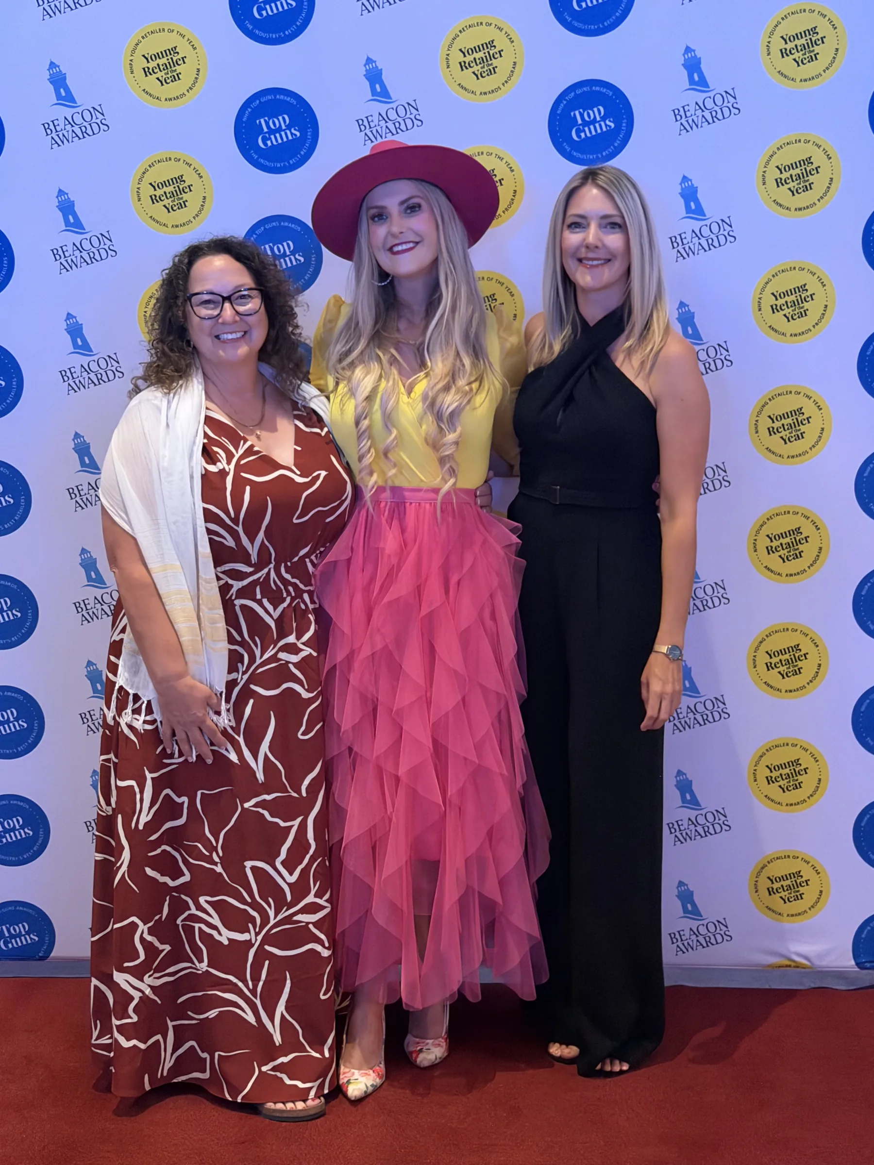 Three women pose on a red carpet before a Beacon Awards, Top Guns and Young Retailer of the Year backdrop.