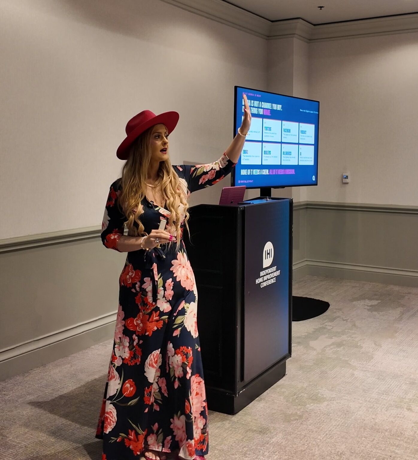 Stefanie Couch, in a pink hat and floral dress, gestures toward a slide beside the IHI podium during The Media Playbook