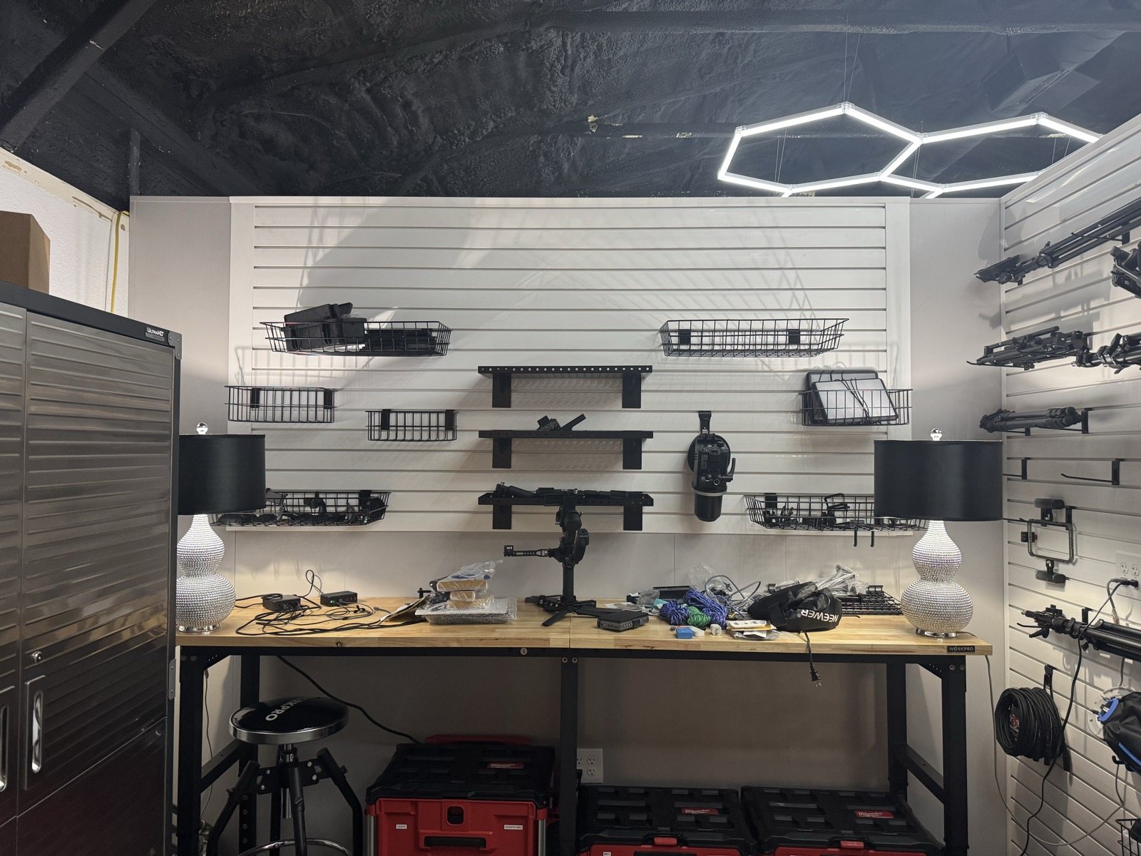 White Trusscore slat wall with shelves and wire baskets above a wooden workbench with two lamps