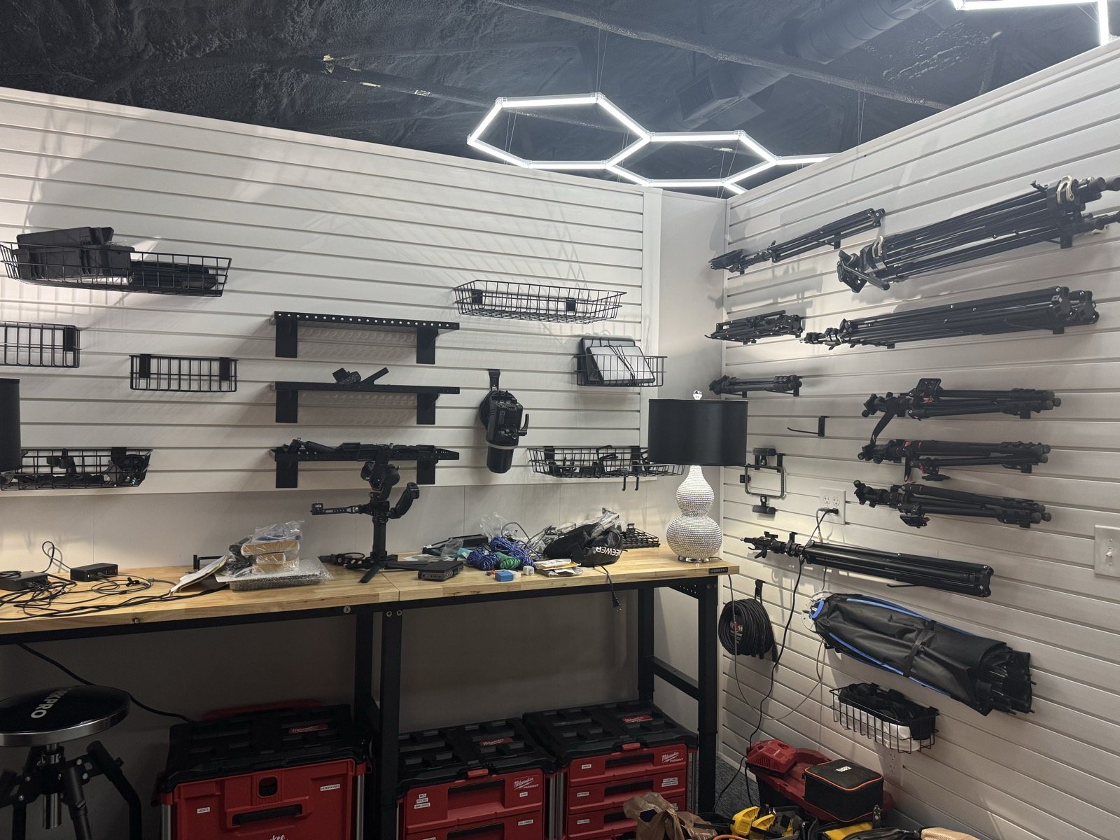Trusscore slat wall with shelves, wire baskets, lights and tripods above a workbench in the Grit Garage