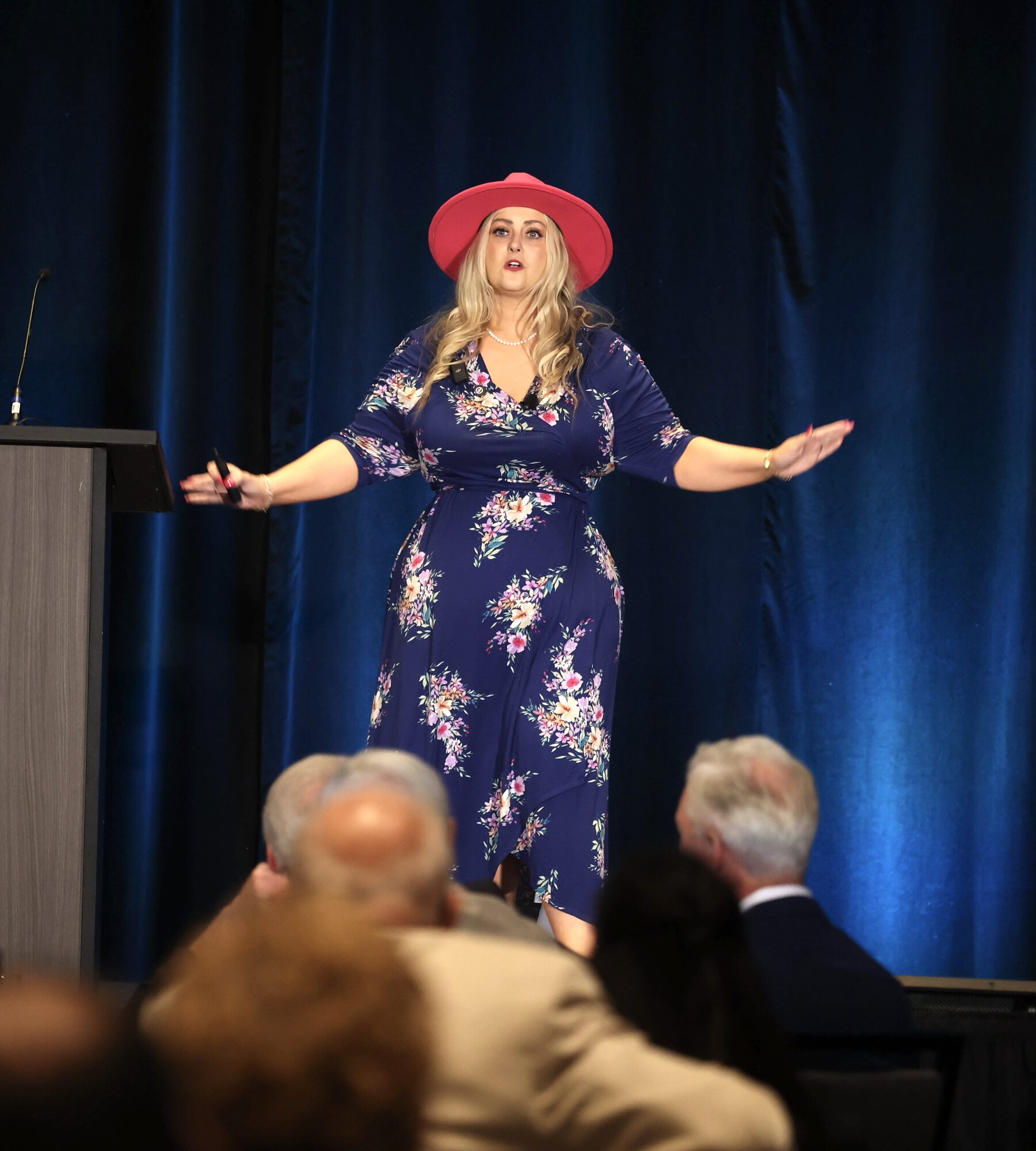 Stefanie Couch in a pink hat and floral dress speaking on stage beside a podium, with seated attendees in the foreground