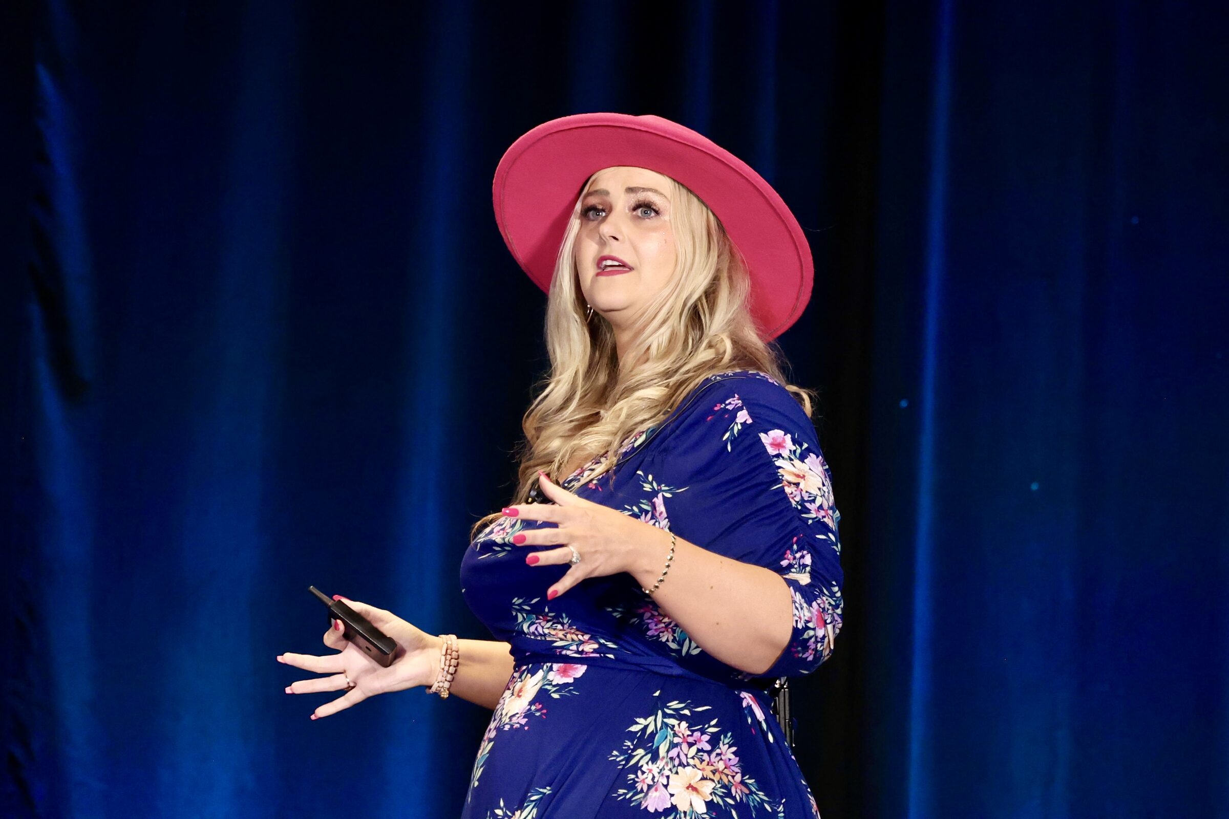 Stefanie Couch holding a presentation clicker and gesturing on stage in front of a blue curtain
