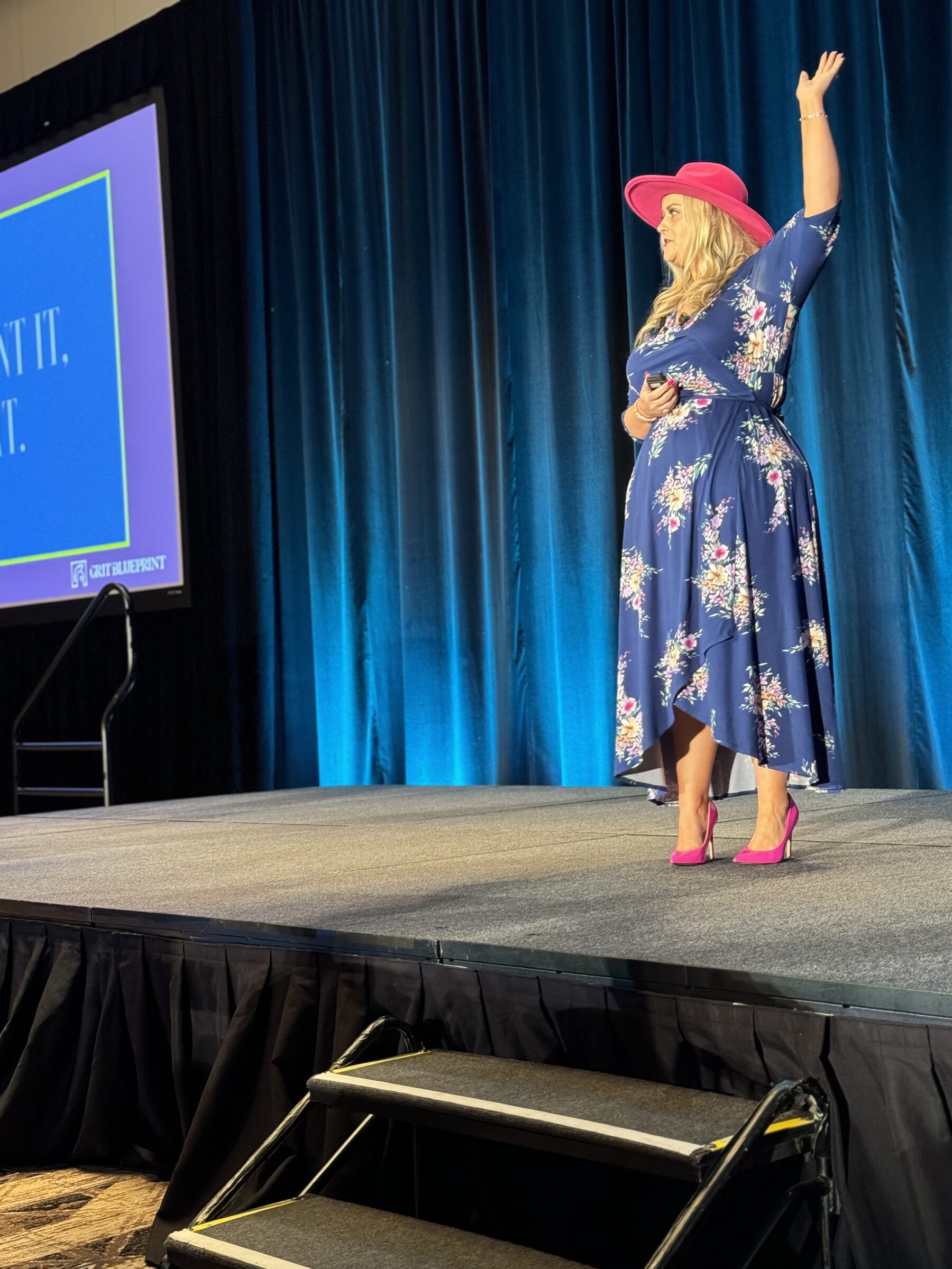 Stefanie Couch on a stage with blue curtains, one arm raised, beside a projection screen