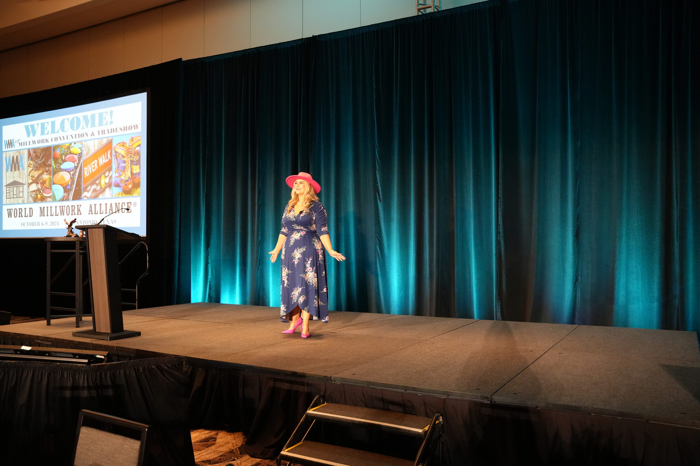 Stefanie Couch on a stage with teal lighting, next to a screen showing a World Millwork Alliance welcome slide
